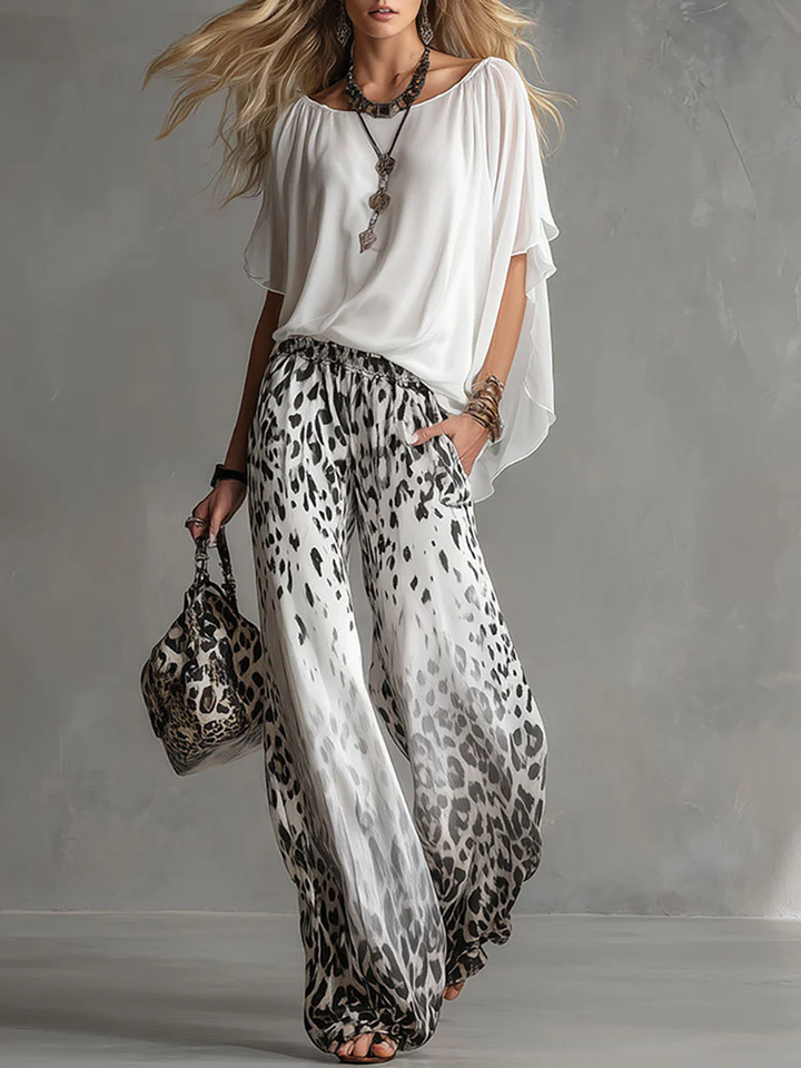Woman wearing a flowing white top and leopard print pants, holding a matching bag in a studio setting.