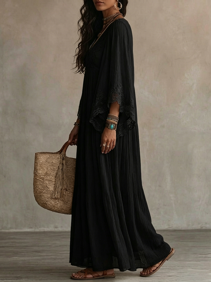 Side view of a model in a black batwing sleeve maxi dress with lace detail, holding a straw bag in a textured indoor setting.