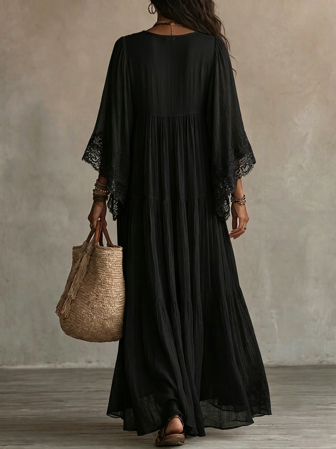 Back view of a black batwing sleeve maxi dress with lace details, worn by a model carrying a straw bag in a simple indoor setting.