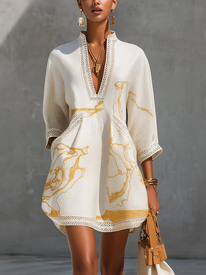 Ivory mini dress with gold motifs and crochet details, V-neckline. Model in jewelry, holding a bag against a concrete wall.