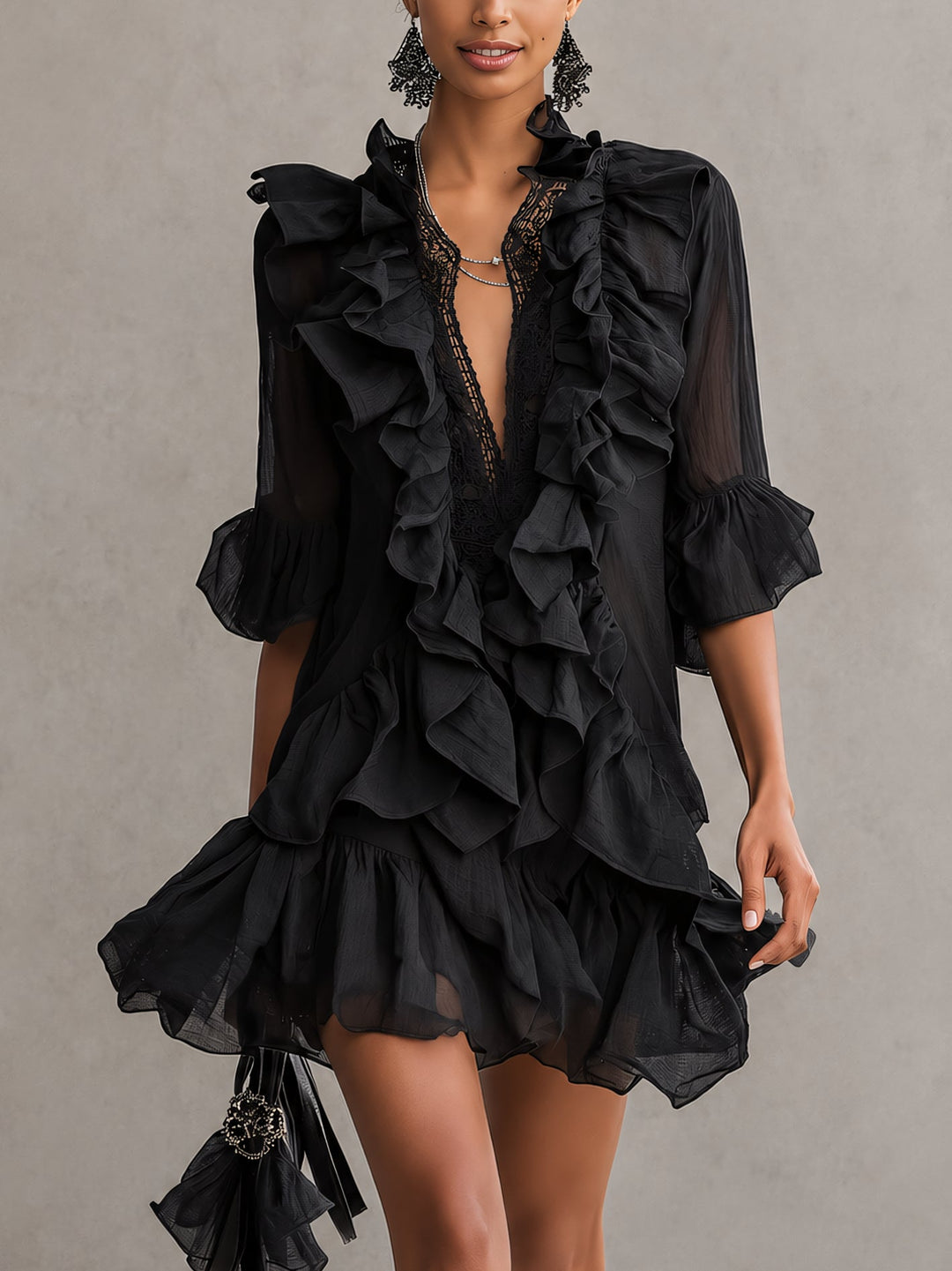 Woman wearing a black mini dress with ruffled layers and lace detail, holding a fringed handbag in a studio setting.