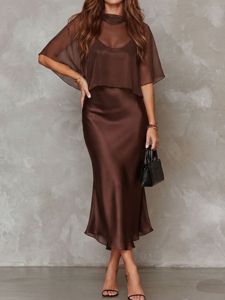 Brown midi dress featuring a sheer overlay, in a studio setting. Model poses with one hand on hip.