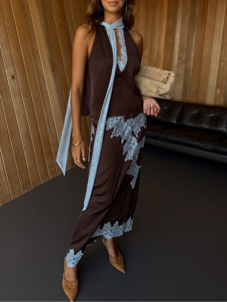 Woman wearing brown lace suit with blue accents, holding beige clutch, standing in wooden room.