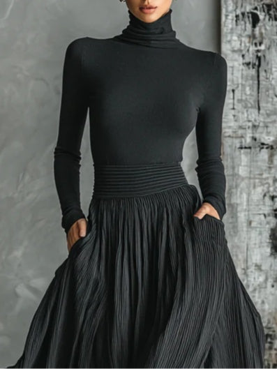 Black turtleneck paired with pleated skirt, hands in pockets. Textured wall in studio setting.