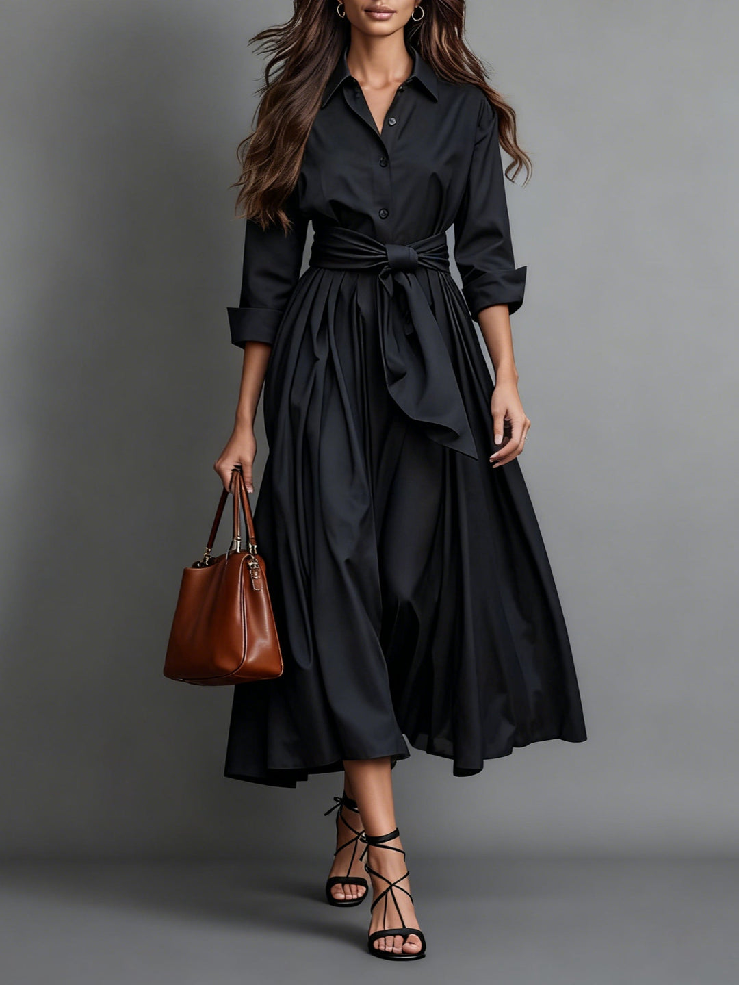 Black maxi dress with waist belt, long sleeves, brown tote bag, and black strappy sandals in studio setting.