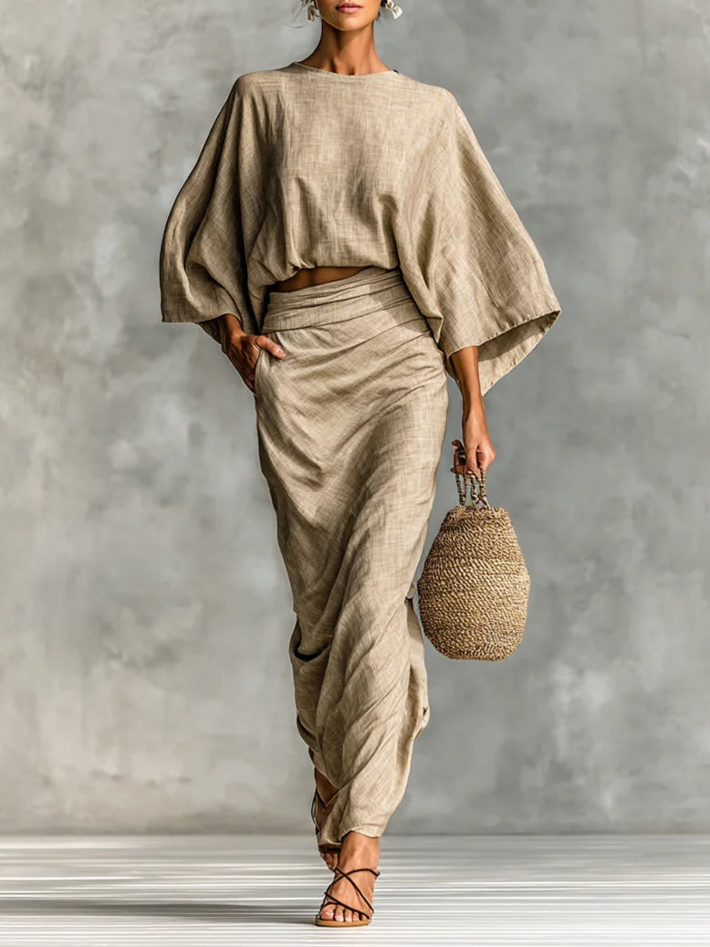 Woman in elegant light brown set with flowy top and draped skirt holding woven bag on striped floor.