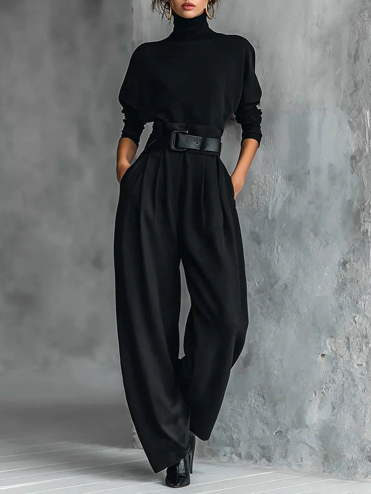 Model in a black elegant top and straight-leg pants set with a belted waist, standing in a textured gray studio.