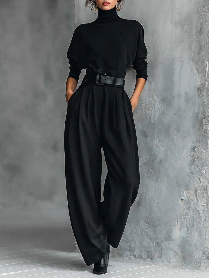 Model in a black elegant top and straight-leg pants set with a belted waist, standing in a textured gray studio.