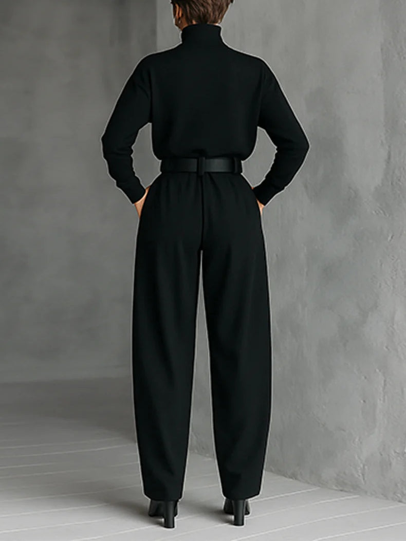 Back view of model wearing a black top and straight-leg pants set with a belted waist in a gray studio.
