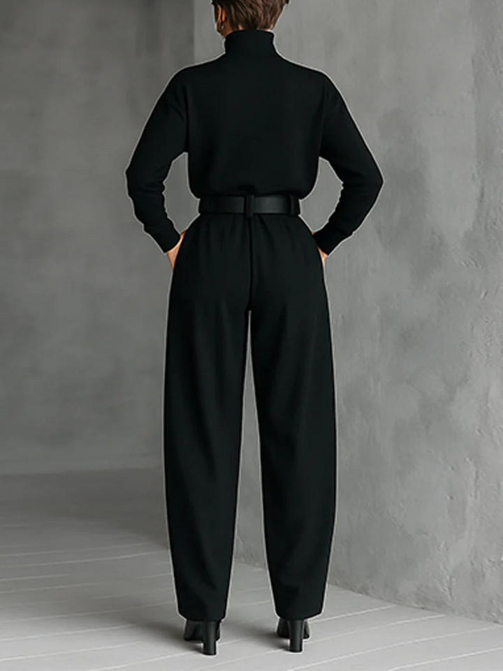 Back view of model wearing a black top and straight-leg pants set with a belted waist in a gray studio.