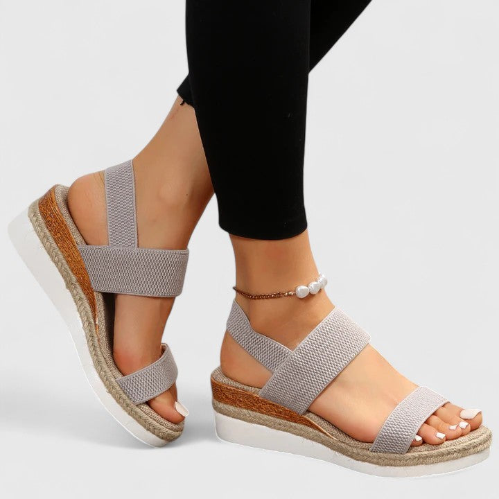Gray orthopedic sandals with supportive straps and platform wedges, worn with black leggings and a pearl anklet in a studio.