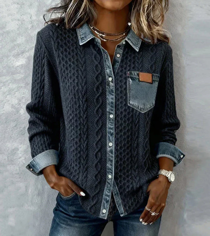 Person wearing a dark knit top with denim accents, posing against a textured gray wall.
