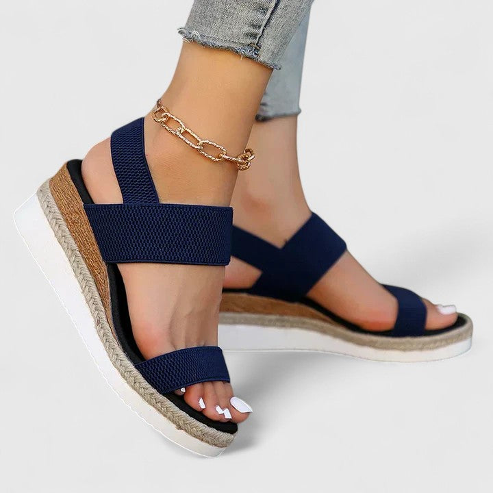 Navy orthopedic sandals with crisscross straps and elevated soles, worn with distressed jeans and a gold anklet in a studio setting.