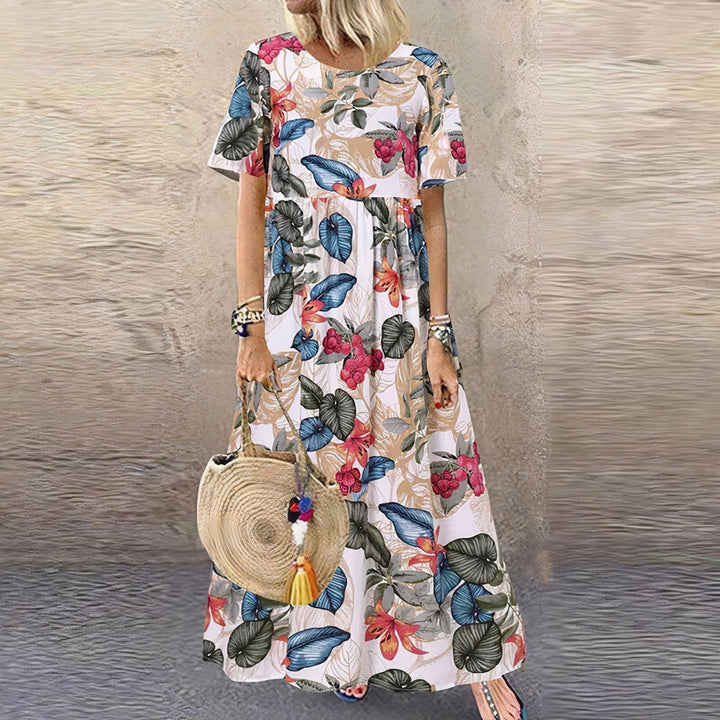 Model wearing a white floral dress with vibrant pink and blue leaves, holding a round woven bag, standing in front of a textured wall.