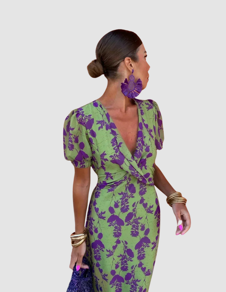 Woman wearing a green dress with purple floral print in a studio setting, accessorized with statement earrings and bracelets.