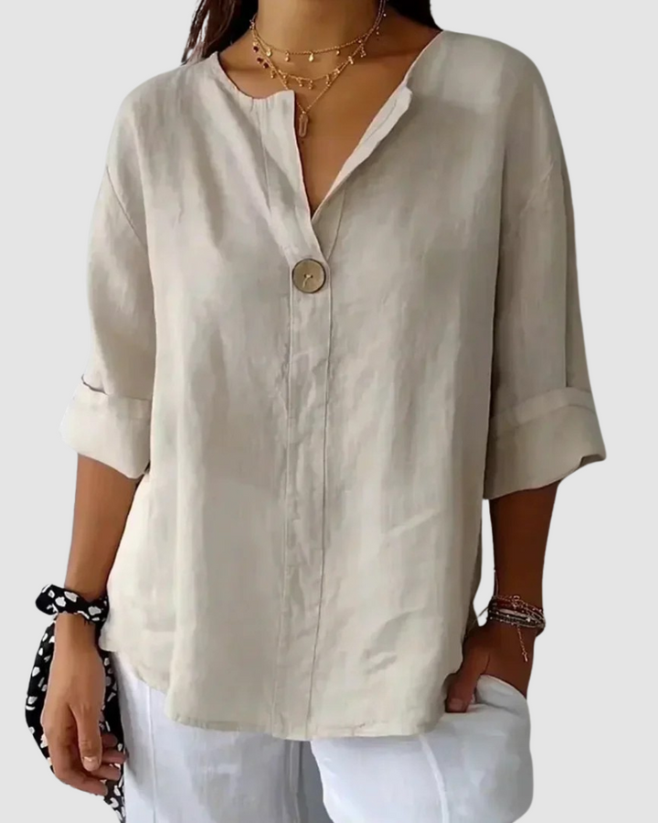Beige blouse with single button front, relaxed fit, folded sleeves, worn with white pants and layered necklaces in studio.