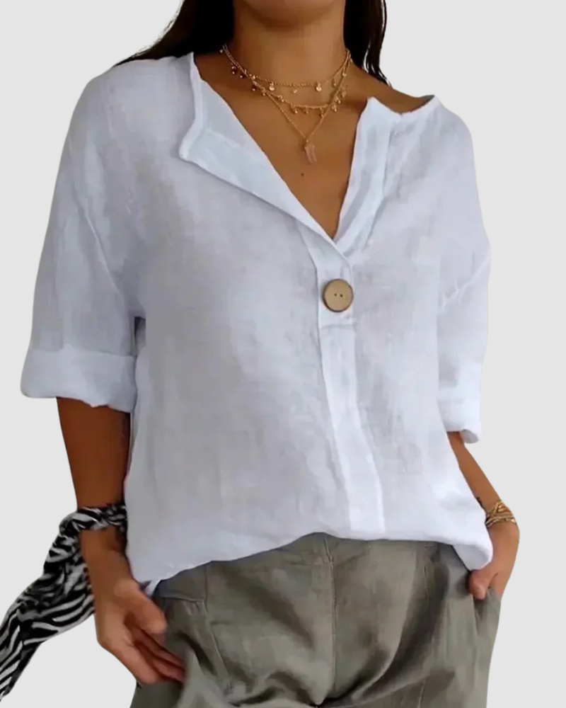 White blouse with button closure, loose fit, rolled sleeves, paired with gray pants, layered necklaces, and black tie in studio.
