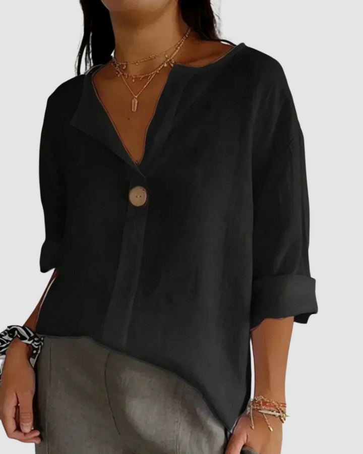 Black blouse featuring single button front, relaxed style, rolled sleeves, paired with gray pants and accessories in studio.