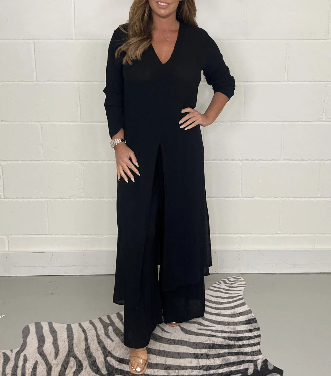Model in a black two-piece set with long sleeves and a V-neck, standing on a zebra-patterned rug against a white brick wall.