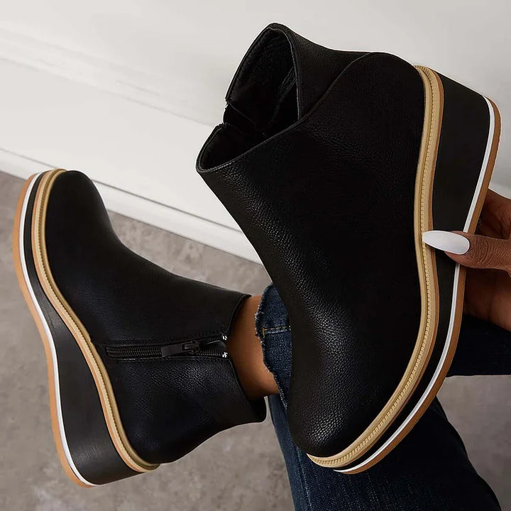 Hand holding a black boot with a wedge sole and zipper against a gray background.