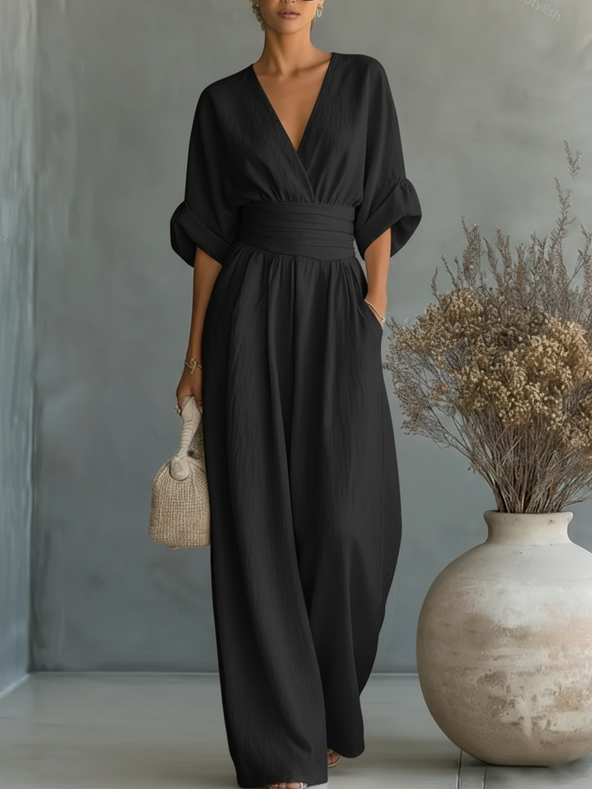 Black V-neck high-waist jumpsuit styled with rolled-up sleeves, modeled in a studio with a large vase and dried plants.