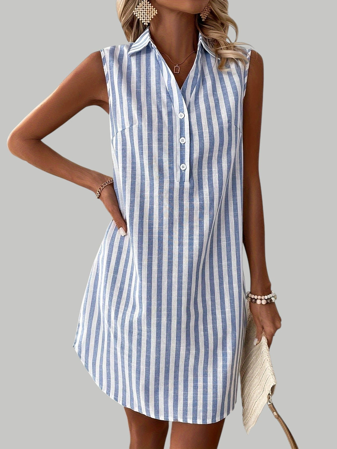 Sleeveless blue striped shirt dress with buttons, worn with bangles and a clutch against a gray studio backdrop.