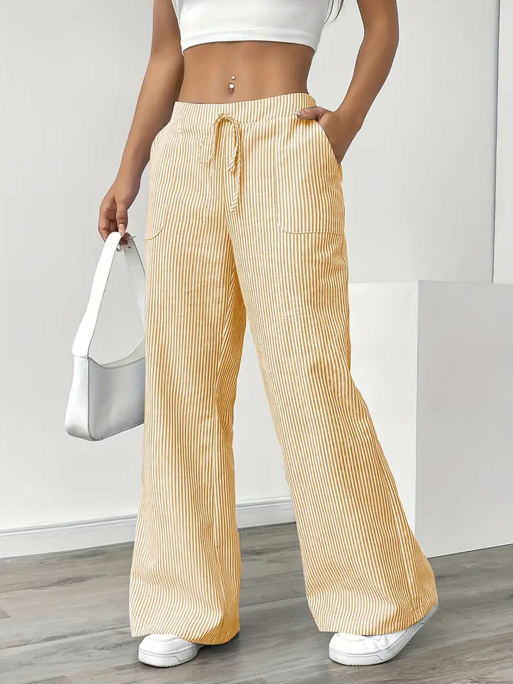 Yellow and white striped pants with pockets and drawstring, modeled indoors with white sneakers and white bag.