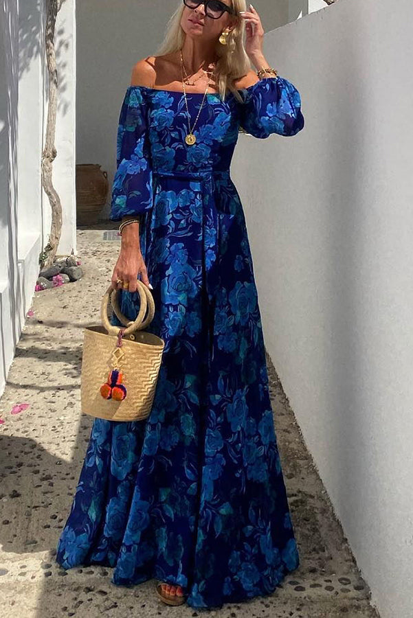 Woman wearing a blue floral off-shoulder maxi dress with long sleeves, holding a straw handbag in a sunlit outdoor setting with white walls.