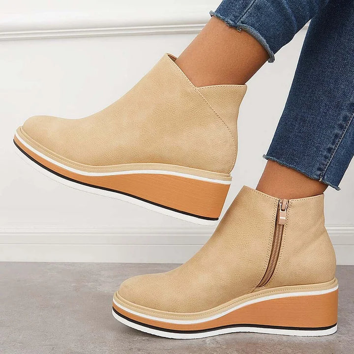 Side view of beige wedge boots with zipper, worn with jeans, against a white wall and gray floor.