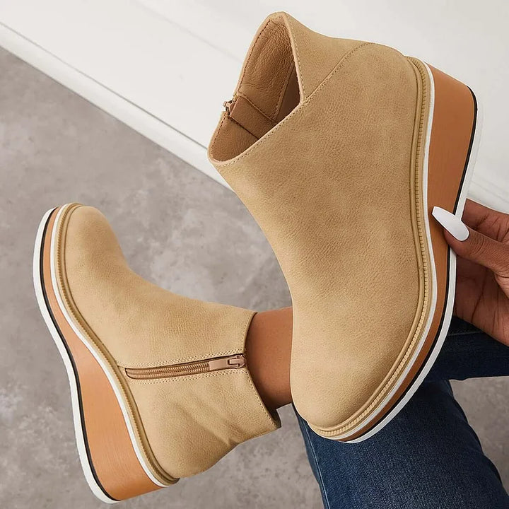 Hand holding a beige boot with a wedge sole and zipper against a gray background.