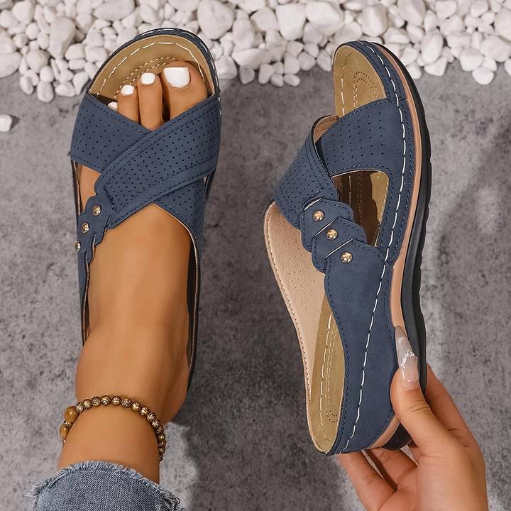Blue orthopedic wedge sandals with perforated straps held in hand, on a textured surface with white stones.