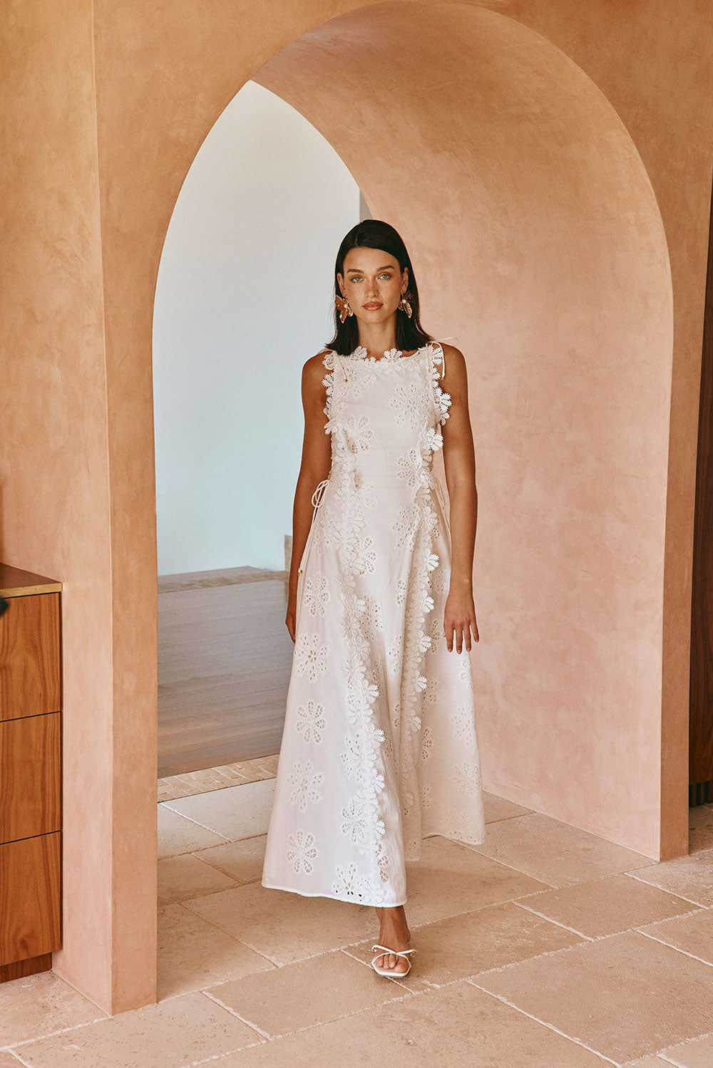 Woman in white floral embroidered maxi dress stands in arched hallway with peach-toned walls.