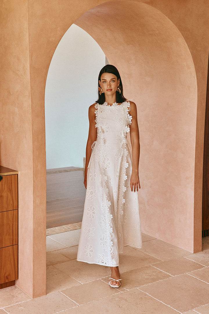 Woman in white floral embroidered maxi dress stands in arched hallway with peach-toned walls.