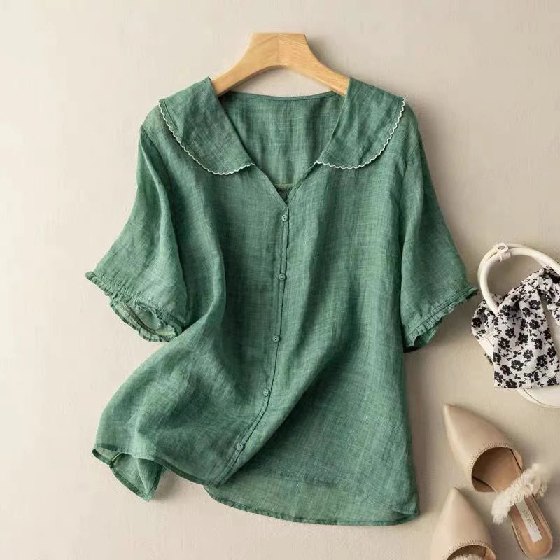 Green short sleeve blouse with delicate trim on hanger, indoor background with shoes and handbag.