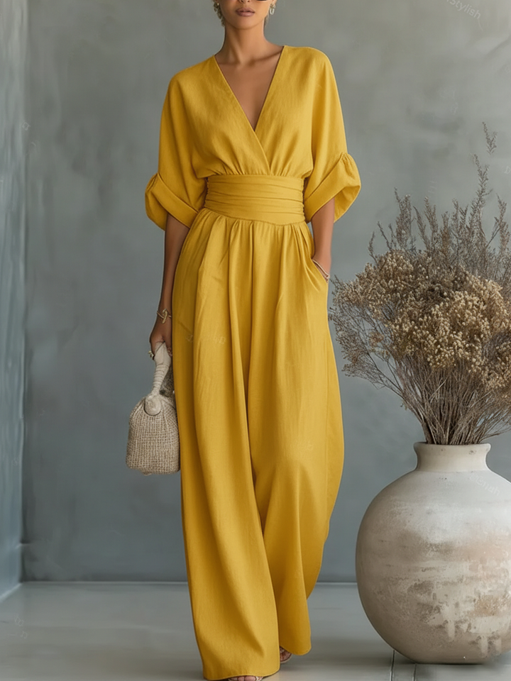 Yellow V-neck high-waist jumpsuit with rolled-up sleeves, displayed in a studio with natural tones and a vase of dried plants.