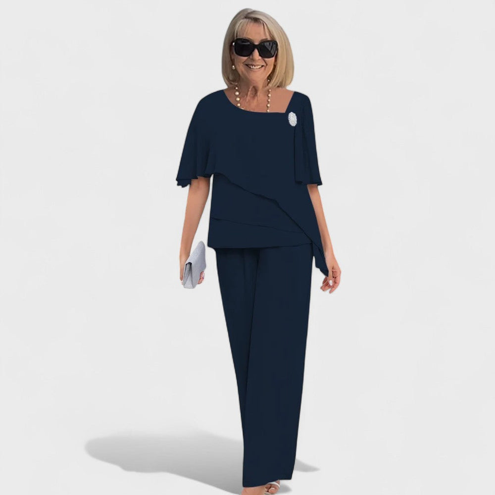 Woman in a dark blue elegant two-piece set with a layered top and wide-leg pants, accessorized with a small clutch in a studio setting.