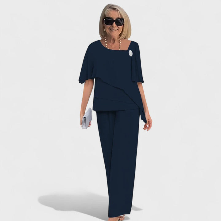 Woman in a dark blue elegant two-piece set with a layered top and wide-leg pants, accessorized with a small clutch in a studio setting.