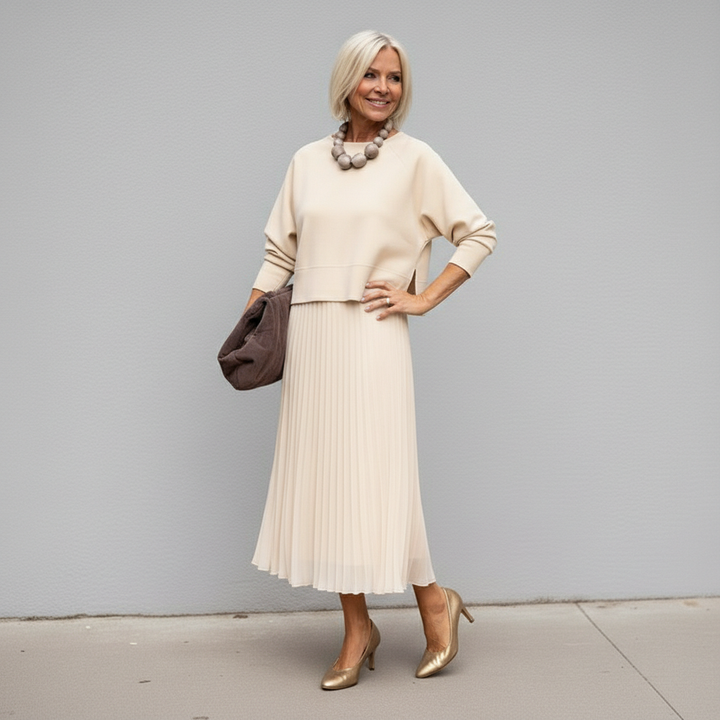 Sophisticated woman in beige outfit with pleated skirt and brown accessory, outdoor setting.