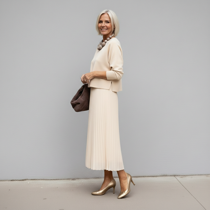 Woman in beige pleated skirt outfit, holding brown clutch, gray wall background.