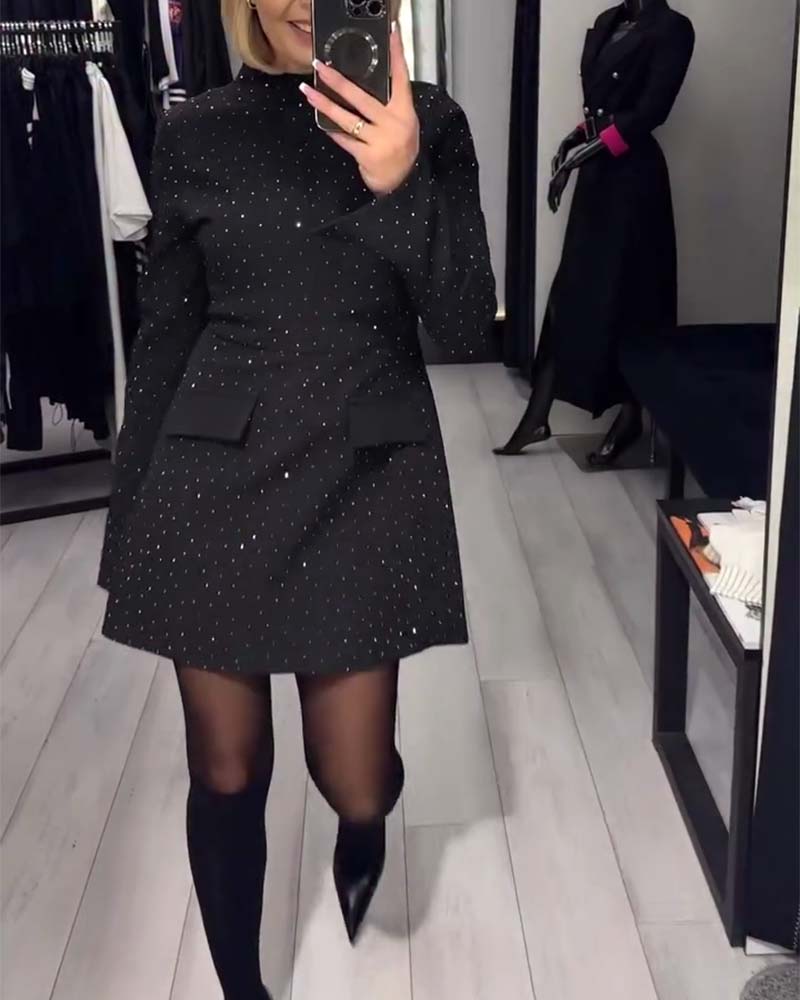 Person modeling a black mini dress with small white dots, long sleeves, and pockets. Displayed in a chic boutique setting with clothing racks and a mannequin.