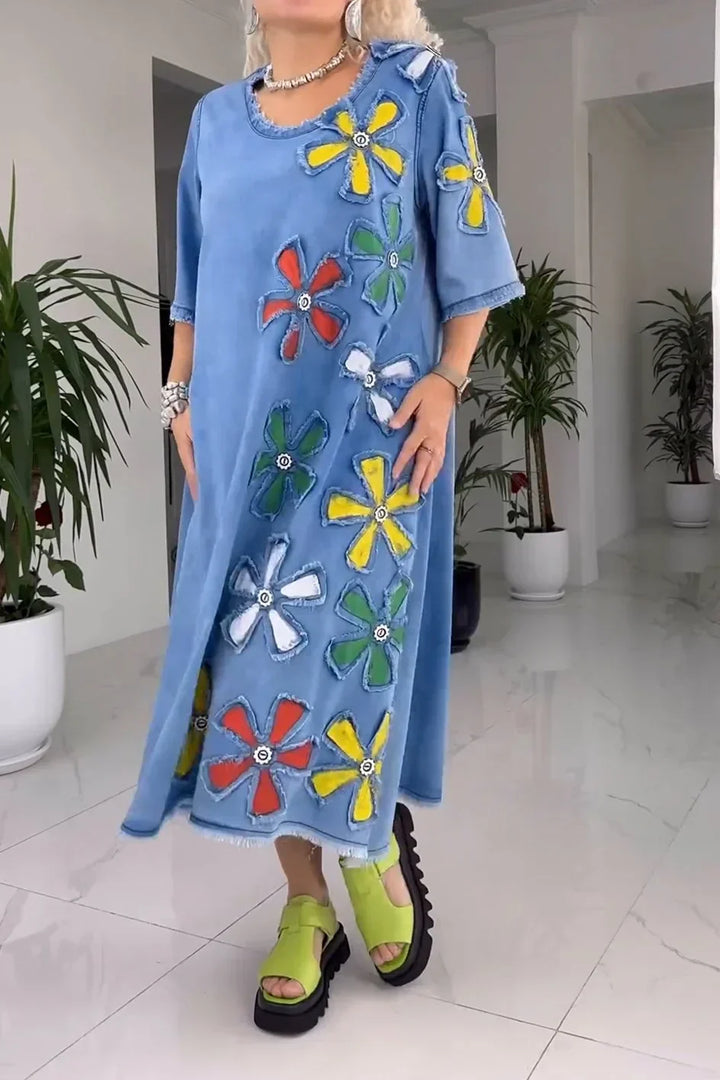 Model in blue floral dress with colorful flower patches, green sandals, in a white room with plants.