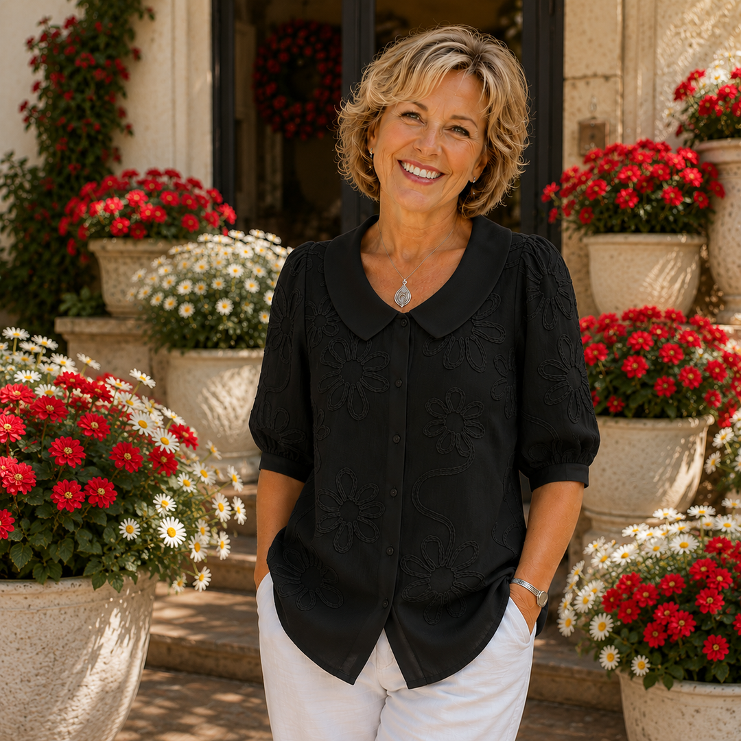 Woman in a black floral blouse poses in garden setting with red and white flowers.