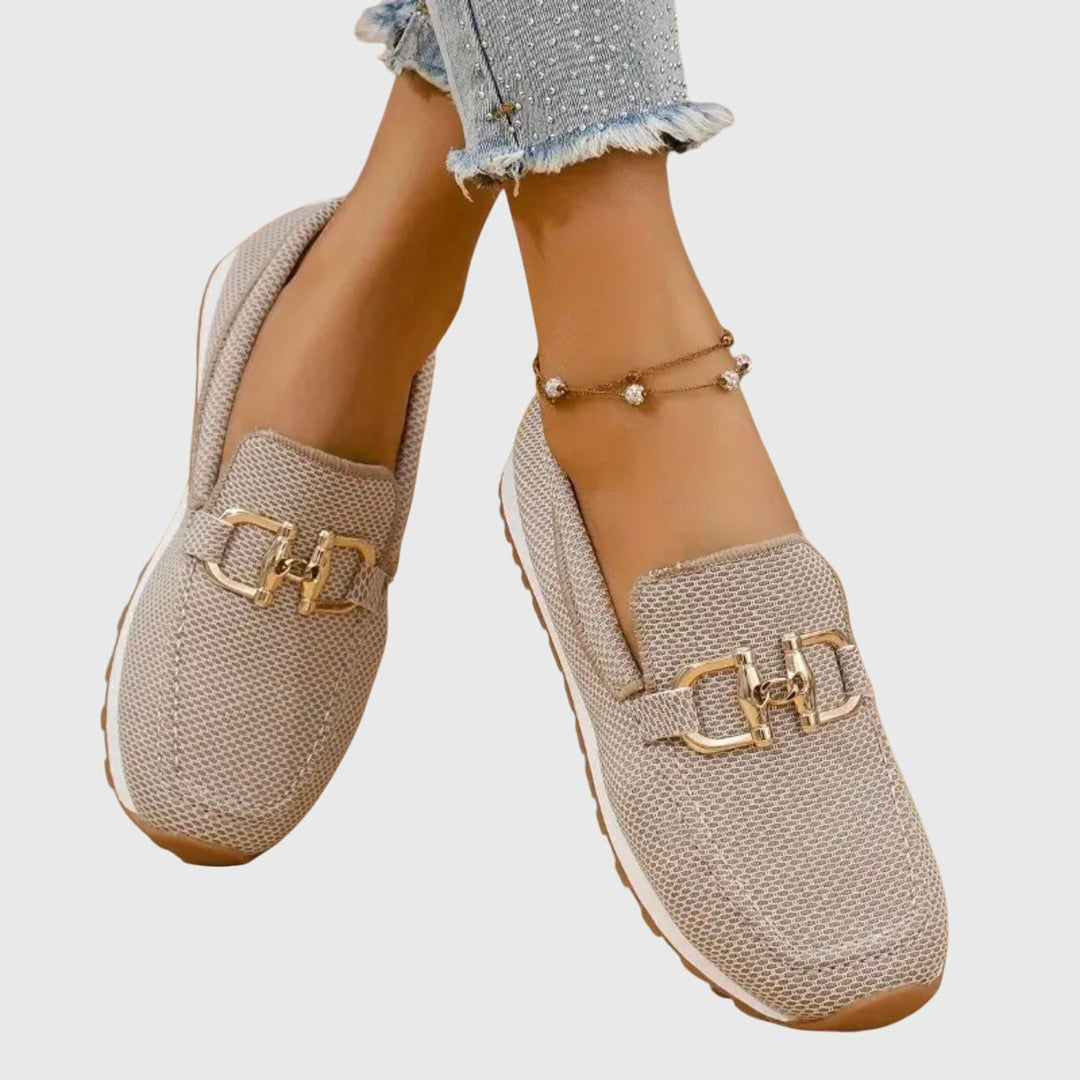 Beige orthopedic loafers with gold accents worn with frayed jeans and anklet on a light background.