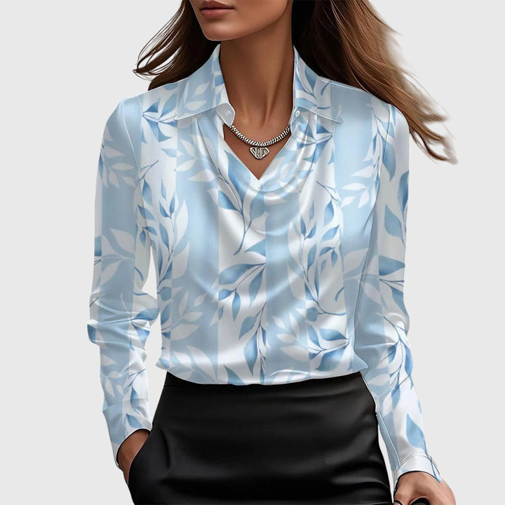 Blue and white floral patterned blouse with long sleeves and draped neckline, worn with a black skirt on a neutral background.