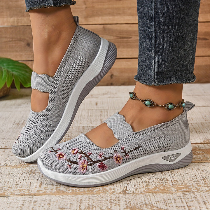 Gray orthopedic shoes with floral embroidery, worn with jeans and anklet, on wooden flooring.