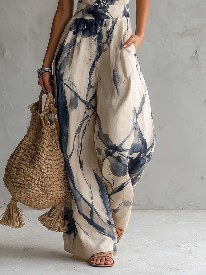 Bottom of flowing jumpsuit in blue abstract pattern on beige, paired with woven bag and sandals. Studio setting.
