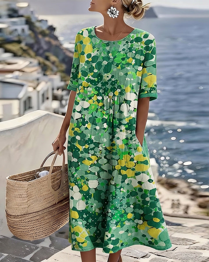 Model in green and yellow floral midi dress, holding bag on terrace with ocean view.