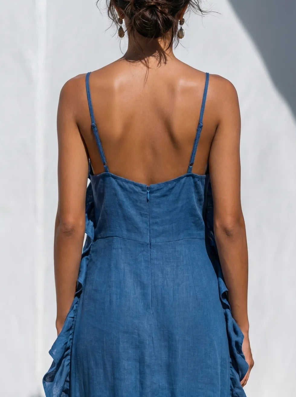 Back view of blue maxi dress with thin straps and ruffles, set against a light, textured wall background.