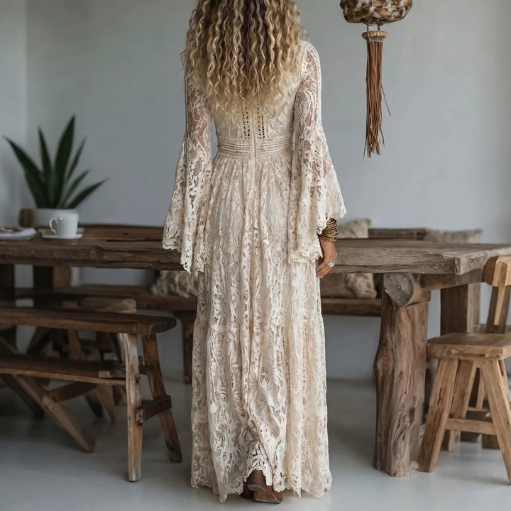 Back view of a woman wearing a cream lace maxi dress, standing in a rustic indoor space with a wooden table and plant.