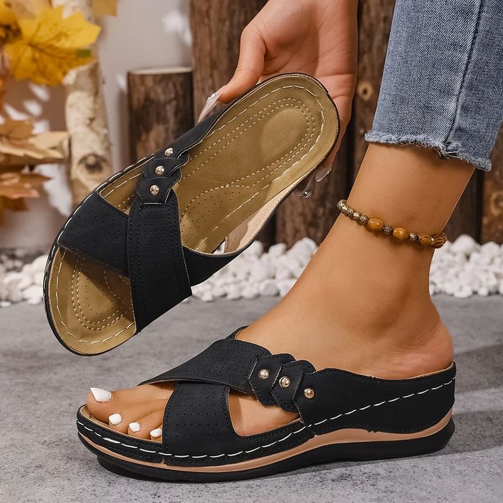 Black orthopedic wedge sandals held in hand, worn indoors with logs and autumn leaves in the setting.